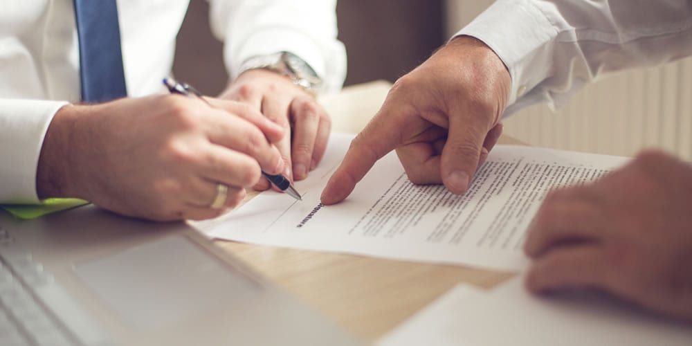 Duas maos sobre a mesa, uma apontando a linha do contrato e outra assinando